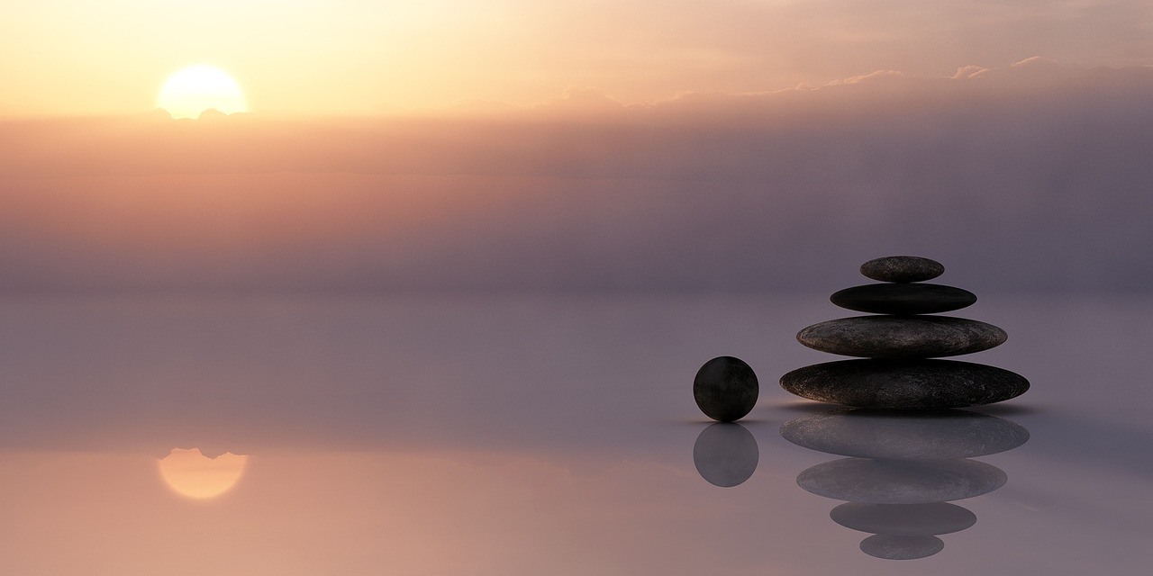 A stack of smooth dark stones rests on still water at sunset, accompanied by a separate round stone and soft reflections.