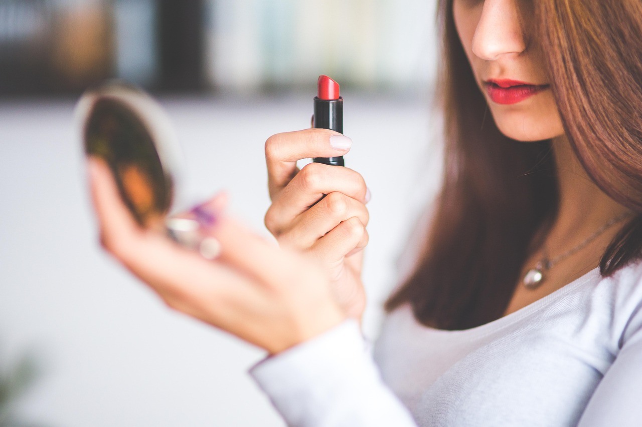 A woman holds red lipstick beside a round compact mirror, with her face and hands softly framed by bright indoor light.