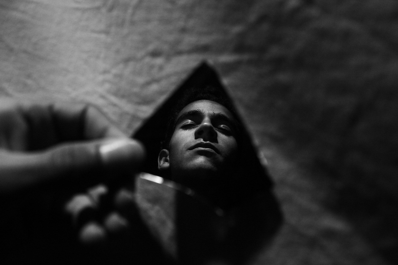 A dark triangular mirror reflects a man’s upturned face while a blurred hand reaches across the foreground.