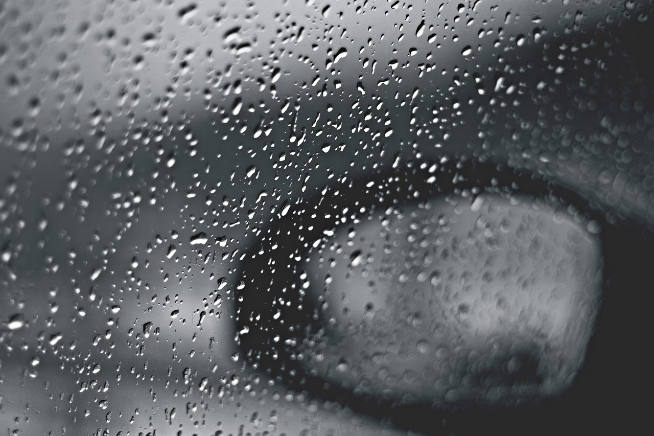 Raindrops cover a window while a car side mirror and pale vehicle shapes blur beyond the wet glass.