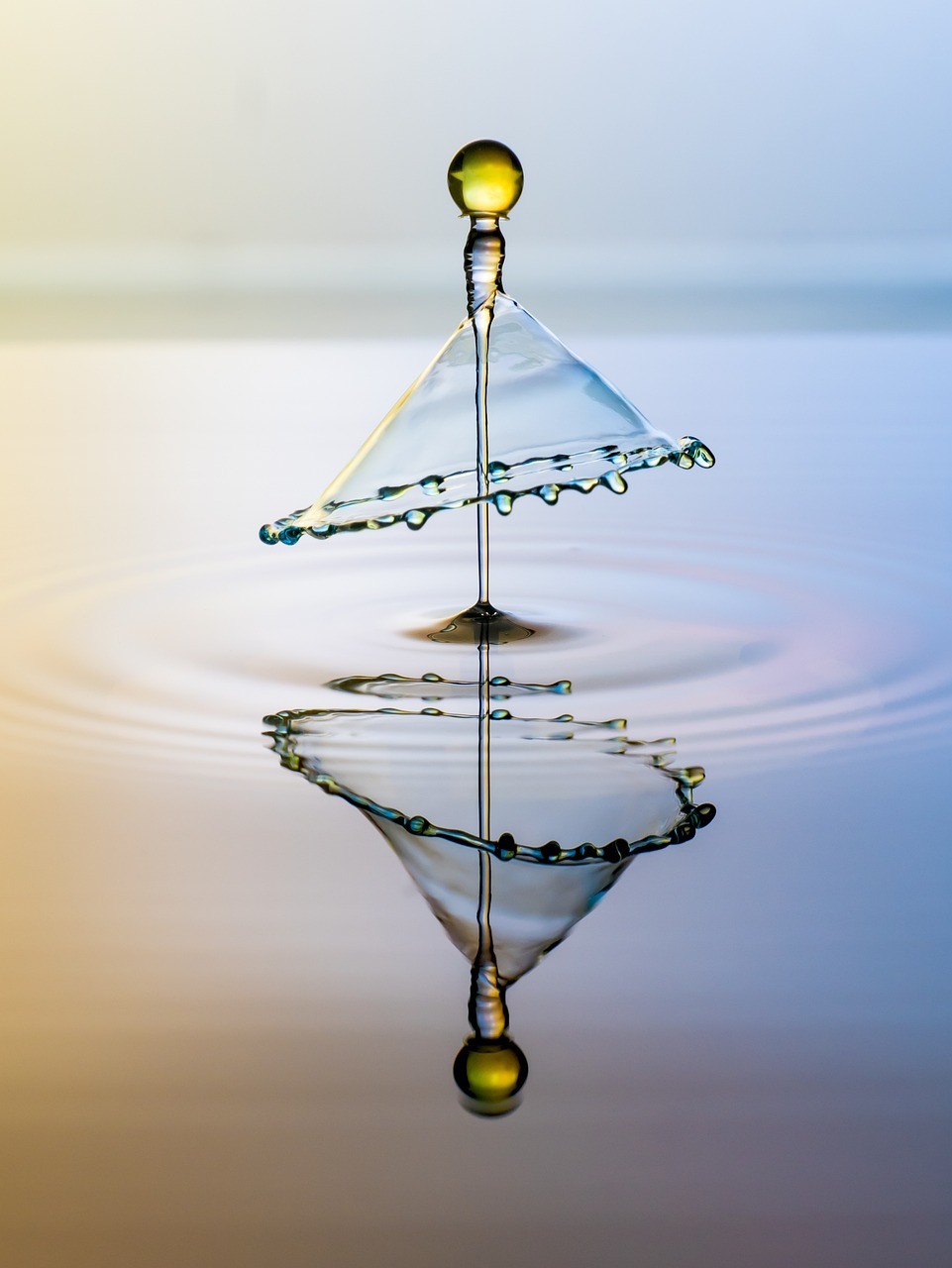A suspended droplet strikes calm water, forming a delicate umbrella-like crown and a vertically aligned reflection below.